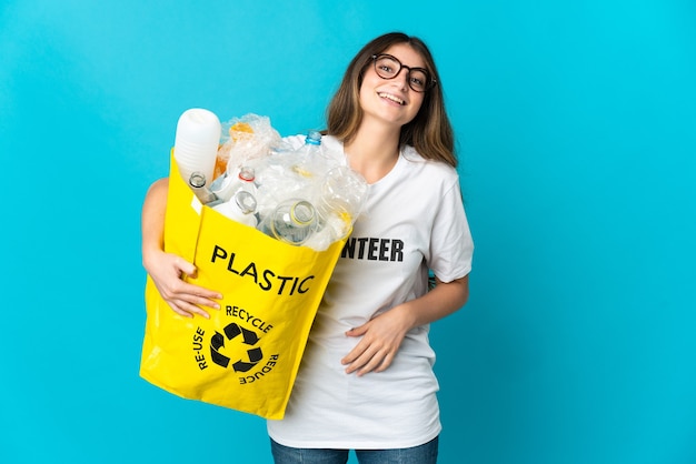 Woman holding a bag full of bottles to recycle isolated on blue smiling a lot