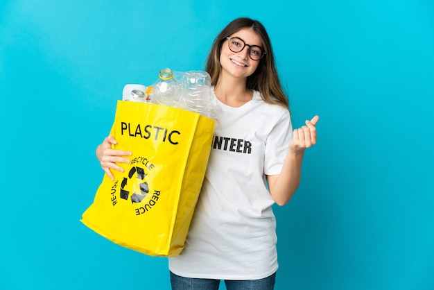 Donna che tiene una borsa piena di bottiglie da riciclare isolata sulla parete blu che fa gesto di soldi