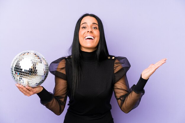 Woman holding a ball party isolated on purple wall receiving a pleasant surprise, excited and raising hands