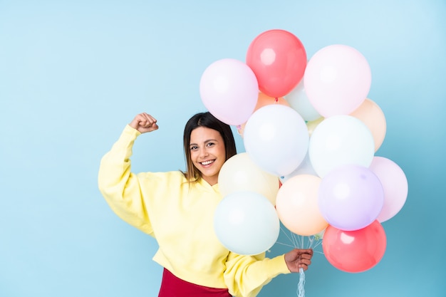 Woman holding balloons in a party over blue wall making strong gesture