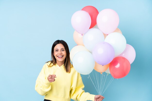 Woman holding balloons in a party over blue wall points finger at you