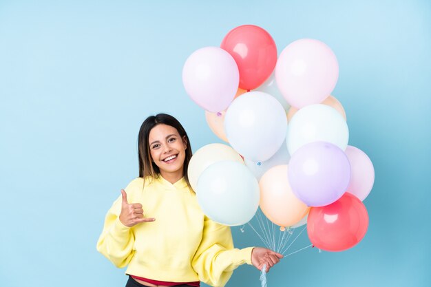 Woman holding balloons in a party over isolated blue wall making phone gesture