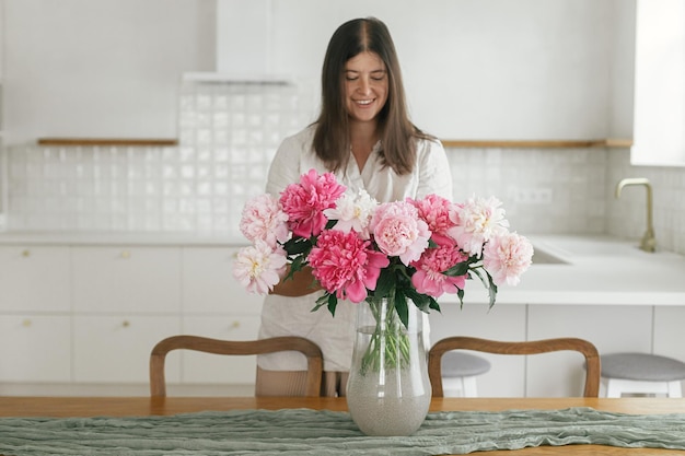 新しいモダンな家の木製テーブルの上の花瓶に美しい牡丹を保持している女性最小限の白いキッチンの引っ越しとハウスキーピングの背景に花でダイニング ルームを飾る若い女性