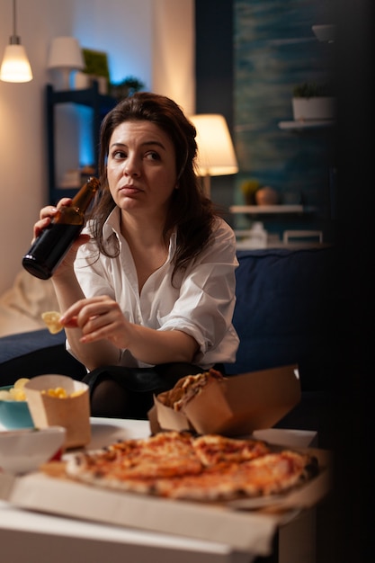 Woman holding beer bottle relaxing on couch in living room watching documentary series