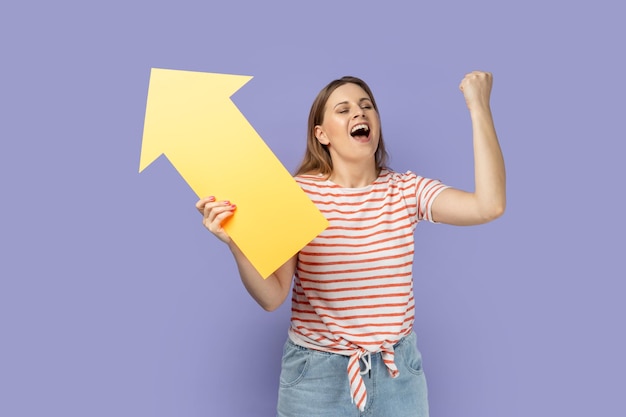 Woman holding big yellow arrow pointing aside and clenched fist celebrating victory increase