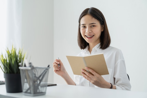 オフィスのテーブルに空白のノートブックとペンを保持している女性。