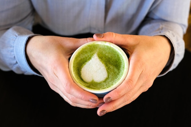 Photo woman holding blue cup with hot trendy green latte with art heart on the foam on black wooden table background healthy drink with latte art