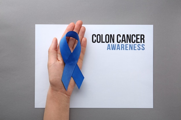 Woman holding blue ribbon near card with words COLON CANCER AWARENESS on grey background top view Space for text