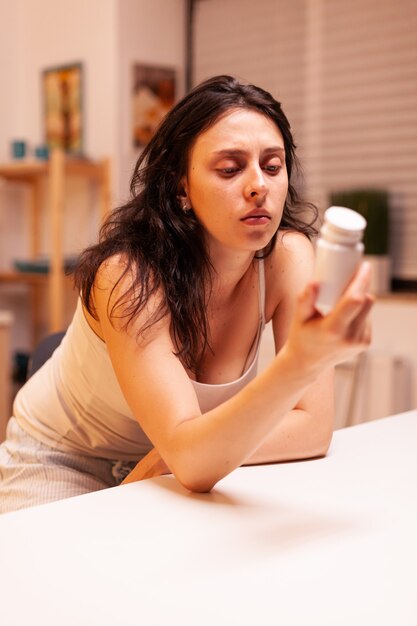 Woman holding bottle with painkillers while having a migraine in home kitchen. worried unwell wife suffering of migraine, depression, disease and anxiety feeling exhausted with dizziness symptoms