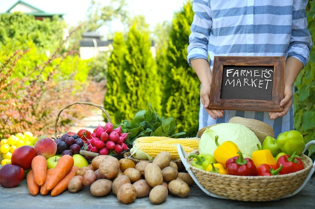 Женщина держит доску возле стола со свежими фруктами и овощами на фермерском рынке