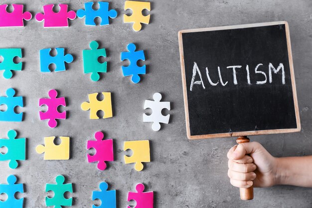 Photo woman holding chalkboard with word autism near colorful pieces of puzzle on grey background