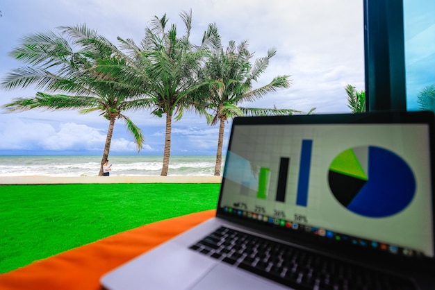 Woman holding coffee cup and smartphone standing under coconut palm tree on sea beach near beachfront house with business graph and charts on laptop blur foreground. Concept of successful life.
