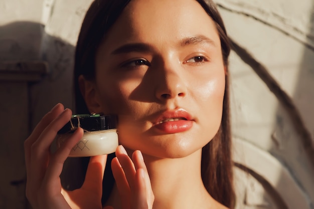 Woman holding cosmetics cream for applying to skin of face. Female hold cream on old wall background with shade from foliage. Advertising concept of healthy lifestyle and self care. Copy space for SPA