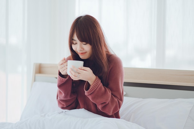 Woman holding a cup coffee tea milk on the bed in a white roomWoman drinking