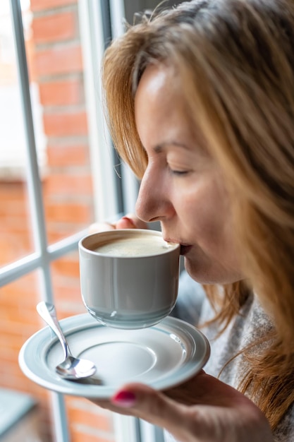 Woman holding a cup of coffee while taking a sip of the drink by the window of her house