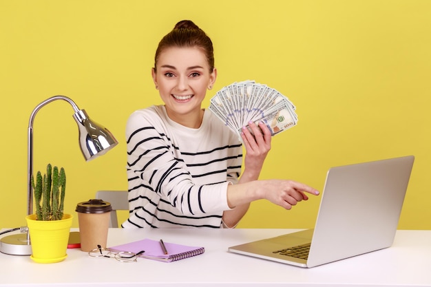 Photo woman holding fan of dollar banknotes and pointing at laptop display looking smiling to camera