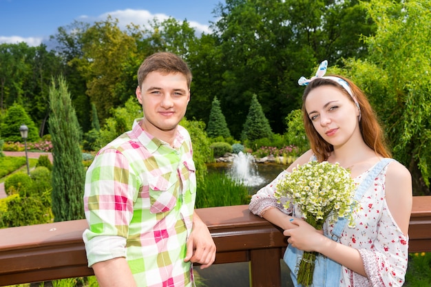 Woman holding flowers while having a date with her  handsome man in green and red plaid shirt standing on a footbridge in the park with background of different trees and fountain