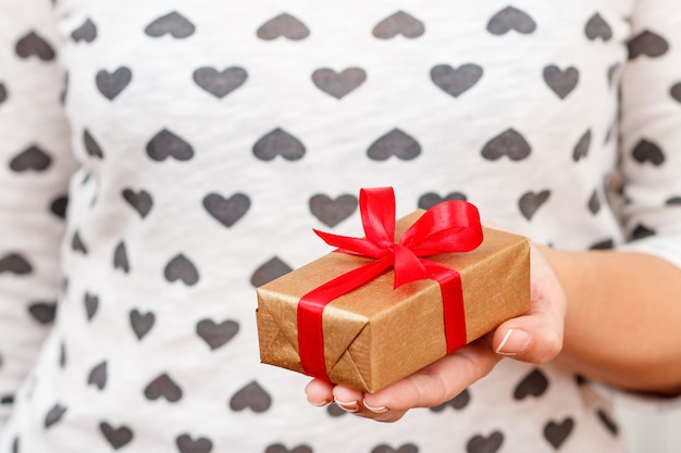 Женщина держит подарочную коробку, перевязанную красной лентой в руках. Малая глубина резкости, выборочный фокус на коробке. Концепция дарения подарка на день святого валентина или день рождения.