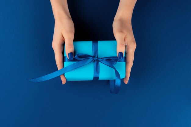 Woman holding gift box with ribbon against classic blue background, top view