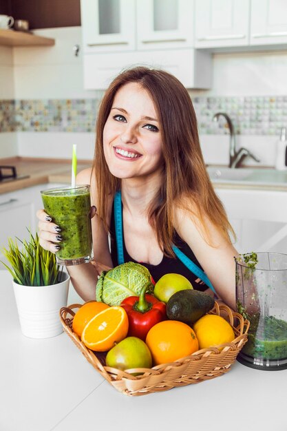 Woman holding glass of green fresh smoothie in the kitchen among tasty fruit and vegetables