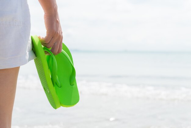 Woman holding green flip flop on sandy beach.