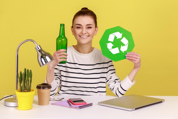 Donna che tiene il segno di riciclaggio verde in mano e una bottiglia di vetro seduta sul posto di lavoro con il computer portatile