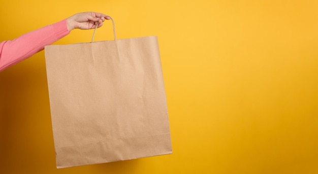 Woman holding in hand brown blank craft paper bag for takeaway
on yellow background. packaging template mock up. delivery service
concept, banner