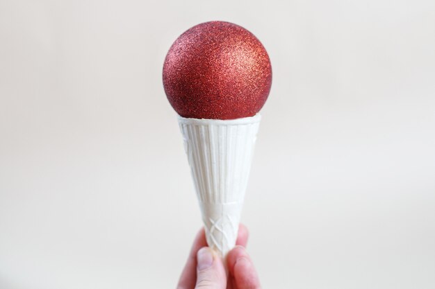 Woman holding in hand ice cream cone with red shiny Christmas ball on beige background