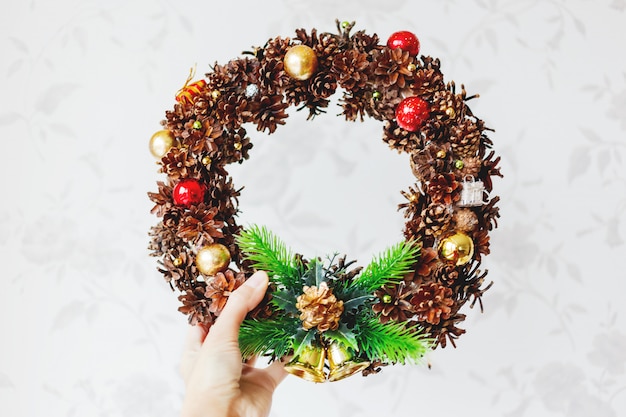 Foto donna che tiene la corona fatta a mano di natale. decorazione fai-da-te di capodanno realizzata con pigne, glitter, sfere e perline decorative, pistola per colla.