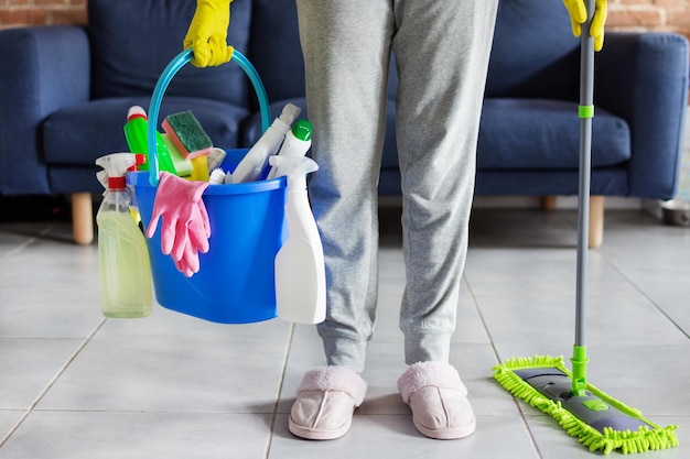 Woman holding in hands bucket with bottles of detergent cleaning equipment and mopping stick in front of sofa in living room Cleaning service idea