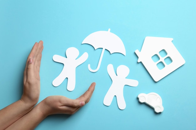 Woman holding hands near paper silhouettes of people with umbrella car and house on color background top view Life insurance concept