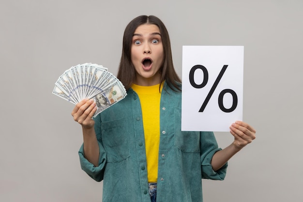 Woman holding in hands percent mark on paper and dollar banknotes looking at camera with open mouth