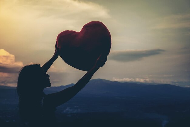 Photo woman holding heart shape against sky during sunset