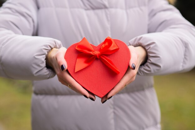 Woman holding a heart shaped red gift box with a bow at the street in the evening