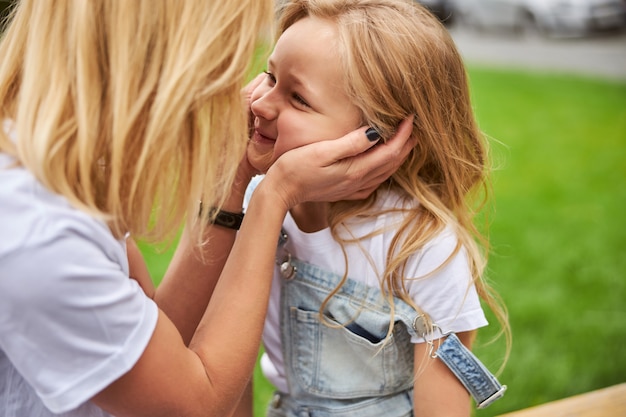 Woman holding her arms on face her daughter while happy kid looking to her mother in the outdoors