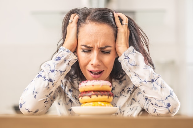 Woman holding her head by hands over irresistable pile of donuts.