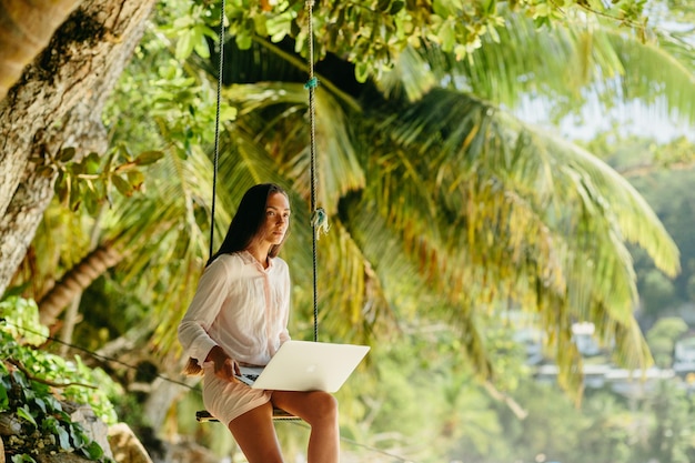 woman holding laptop while sitting by palm trees on swing