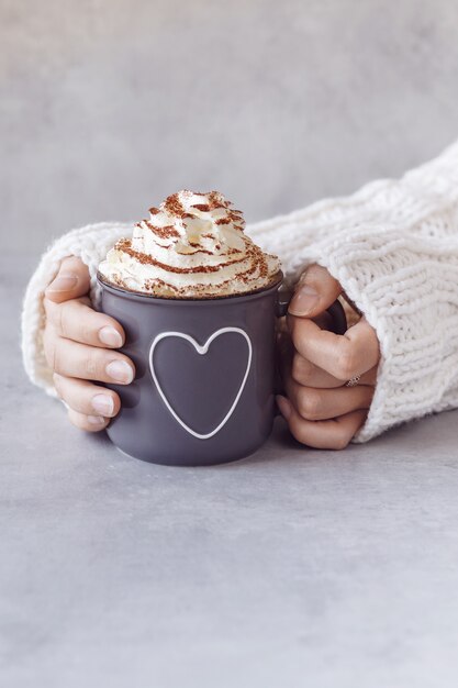 Woman holding metal grey mug of hot chocolate with whipped cream in hands. Grey stone background copy space