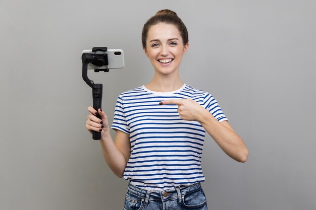 Photo woman holding mobile phone pointing at steadicam looking at camera with positive expression