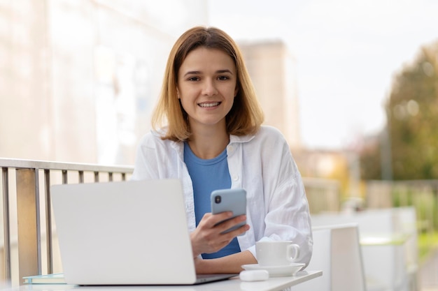 Женщина, держащая мобильный телефон с помощью ноутбука, смотрит на камеру онлайн-общения, сидя в кафе