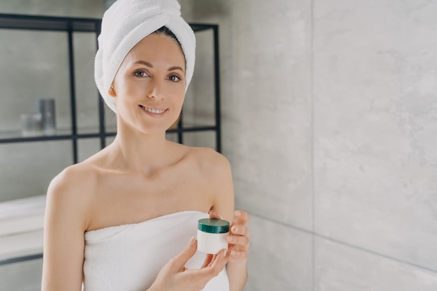 Woman holding mockup cosmetics jar with cream advertising skincare beauty product in bathroom