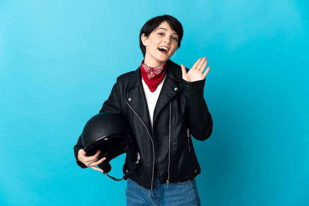 Woman holding a motorcycle helmet on blue saluting with hand with happy expression