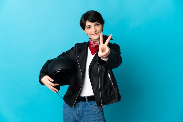 Woman holding a motorcycle helmet isolated on blue background smiling and showing victory sign