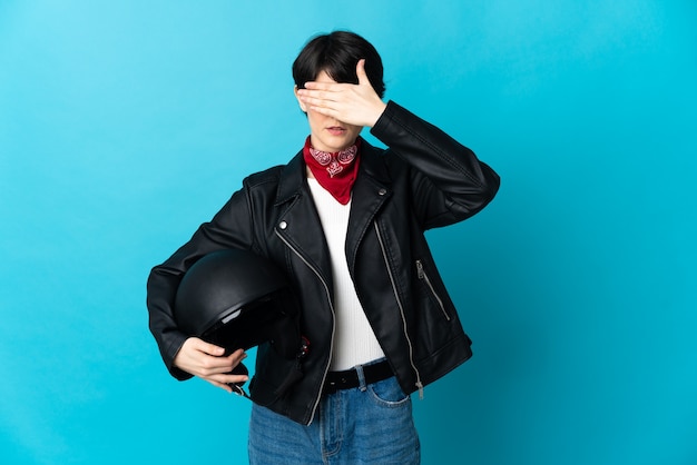 Woman holding a motorcycle helmet isolated on blue wall covering eyes by hands.