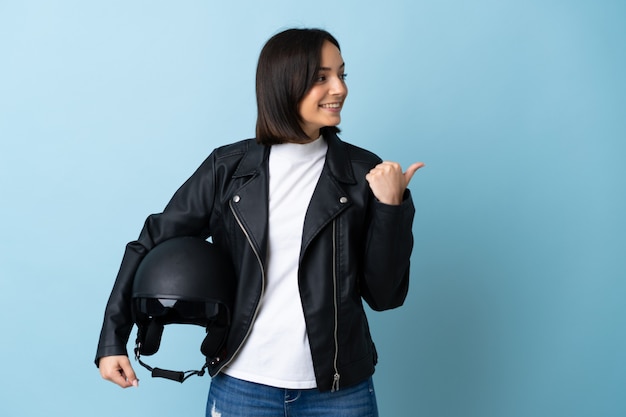 Woman holding a motorcycle helmet isolated on blue wall pointing to the side to present a product