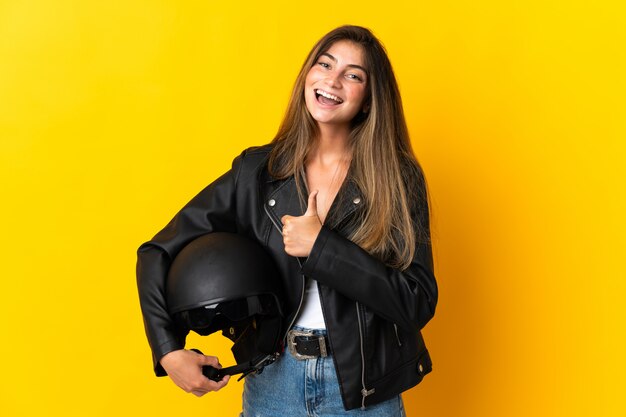 親指のジェスチャーを与える黄色に分離されたオートバイのヘルメットを保持している女性