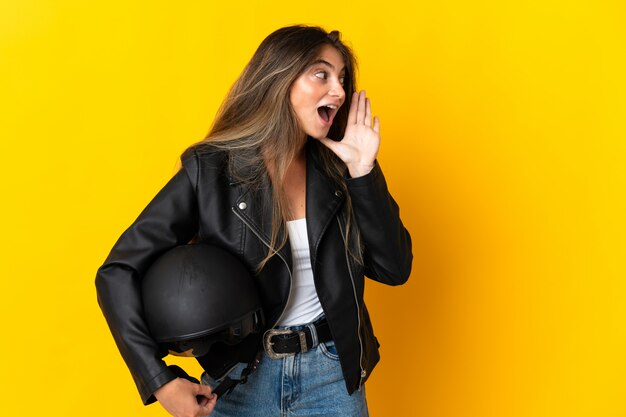 Woman holding a motorcycle helmet isolated on yellow shouting with mouth wide open to the side