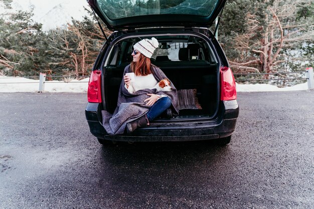 woman holding mug sitting with dog in car trunk