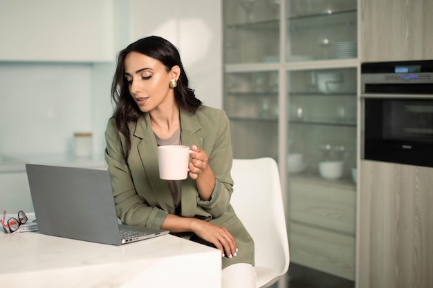 Woman holding mug using laptop at apartment kitchen