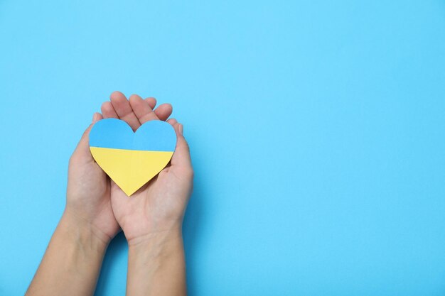 Woman holding paper heart in colors of Ukrainian flag on light blue background top view Space for text